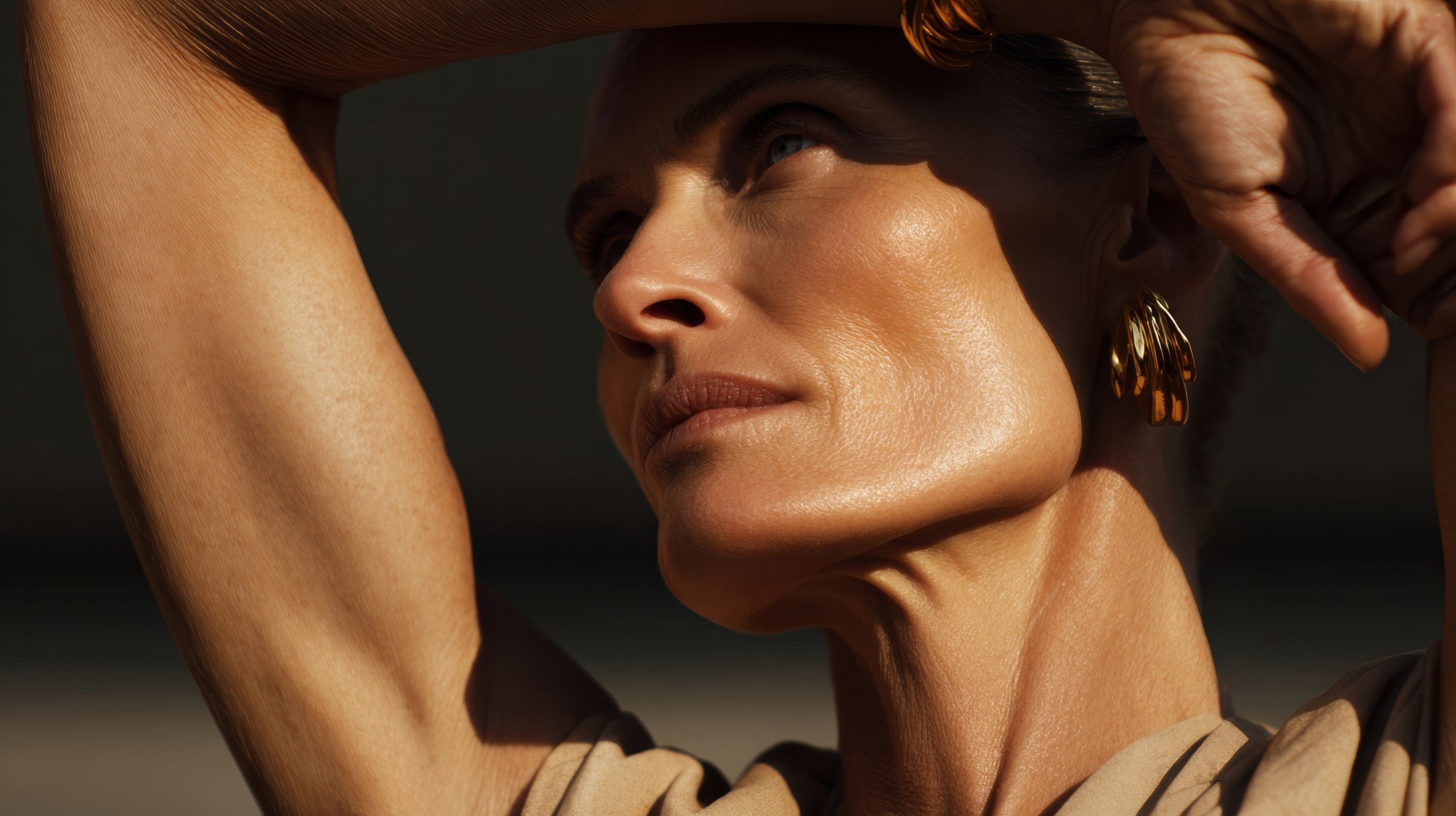 A woman with her arms raised above her head in low golden sun, every line of her face and neck visible, gold sculpted earrings catching the light